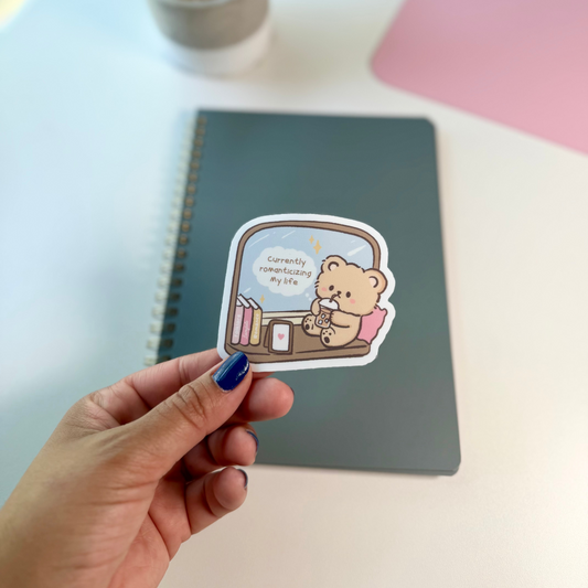 A hand with blue nail polish holds a die-cut sticker featuring a cute cartoon bear sitting in a chair by a window, reading a book. The sticker is shown in front of a dark gray spiral notebook on a white desk, with a potted plant and a pink surface blurred in the background.