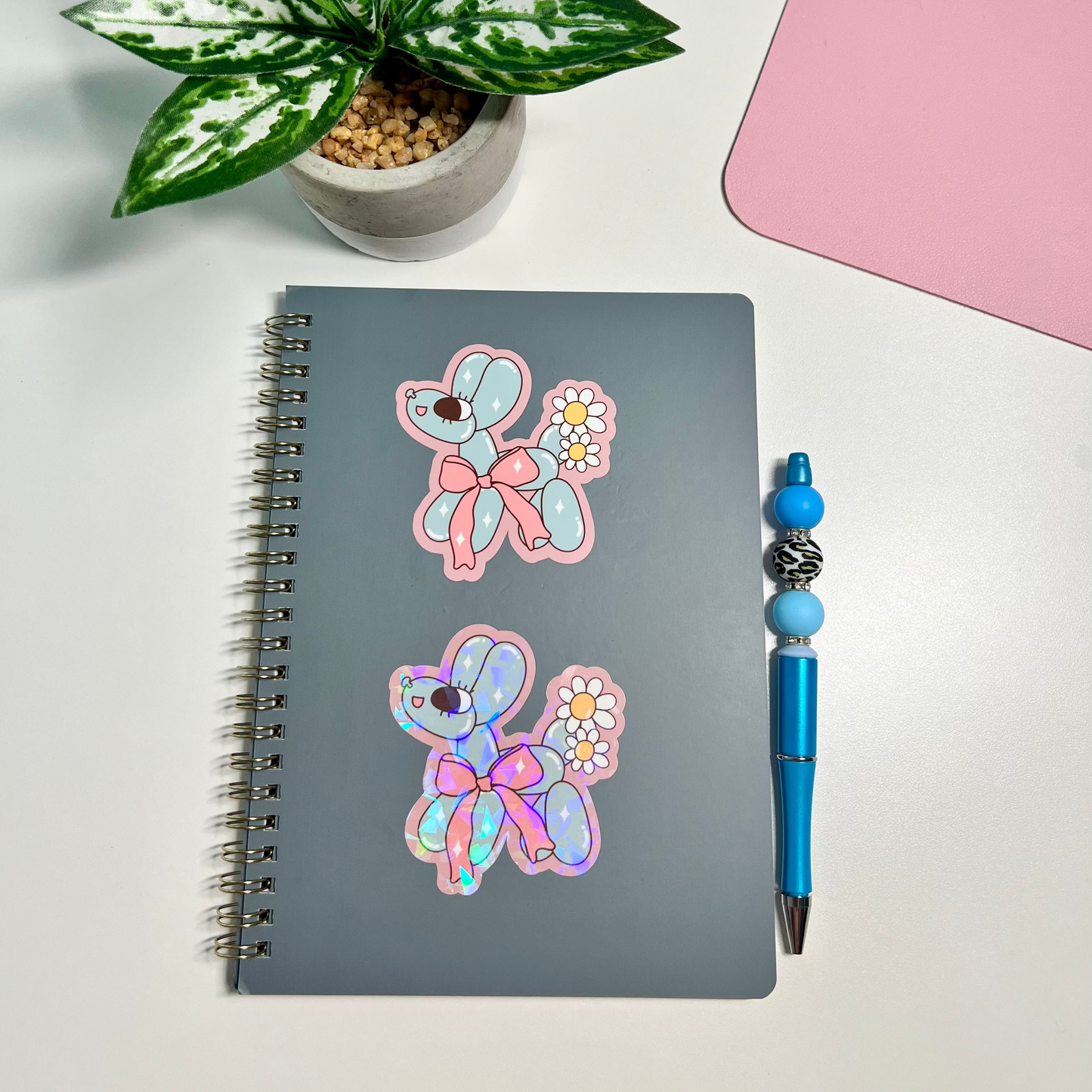 A gray spiral notebook with two iridescent butterfly stickers on the cover, styled on a white desk next to a blue pen and a potted plant.