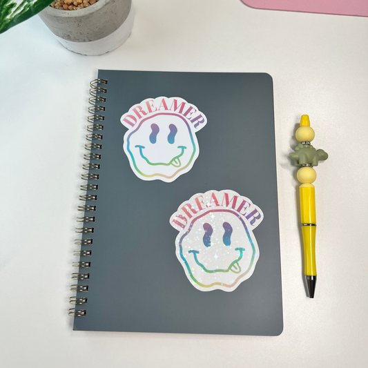 Notebook with 'DREAMER' stickers in glossy and holographic finish , a plant, and a pen on a white surface