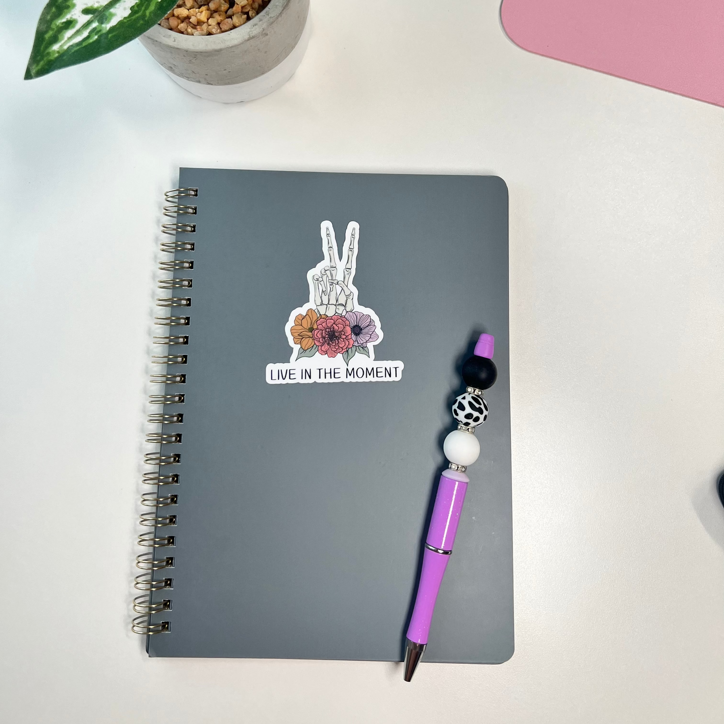 Gray notebook with a peace sign sticker, purple pen, and potted plant on a white surface