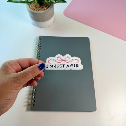 A hand with blue nail polish holds a white sticker that reads 'I'M JUST A GIRL' with a pink bow graphic, placed on a gray spiral notebook on a white desk. A small potted plant and a pink laptop are in the background.