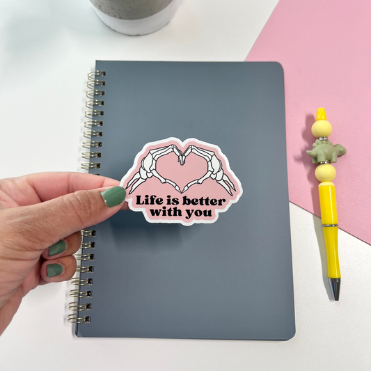 Notebook with a 'Life is better with you' sticker, hand holding the sticker, plant and pen in the background.