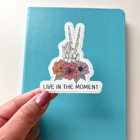 A skeleton hand showing a peace sign with flowers on it's wrist area and the quote LIVE IN THE MOMENT