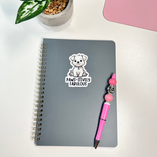 Gray notebook with a sticker of a bear and text, pink pen, and potted plant on a white surface