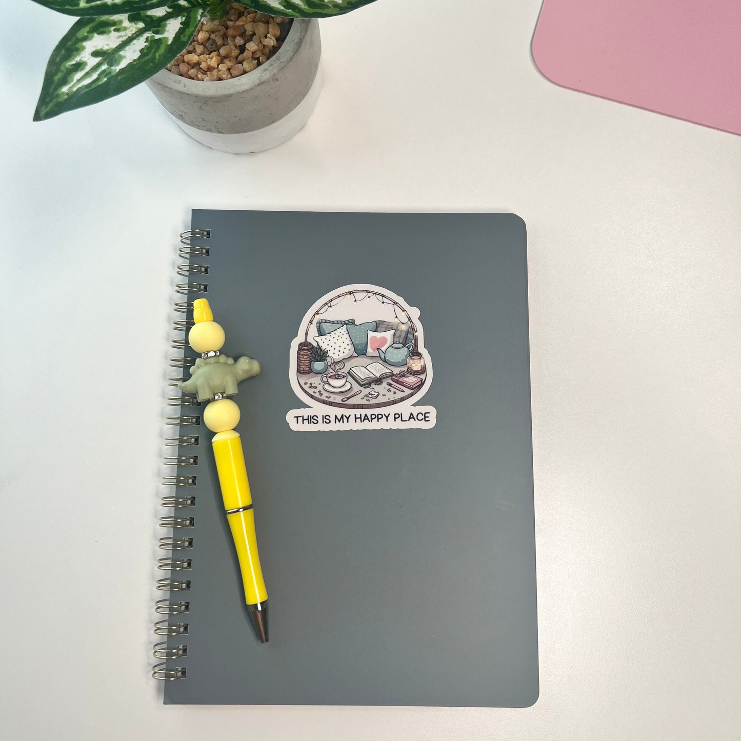Notebook with a yellow pen and decorative sticker on a white surface with a plant in the background