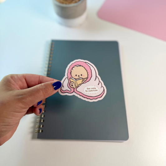 A hand with dark blue nail polish holds a die-cut sticker featuring a cute cartoon bear character wrapped in pink and white comforter, positioned over a gray spiral notebook on a white desk.