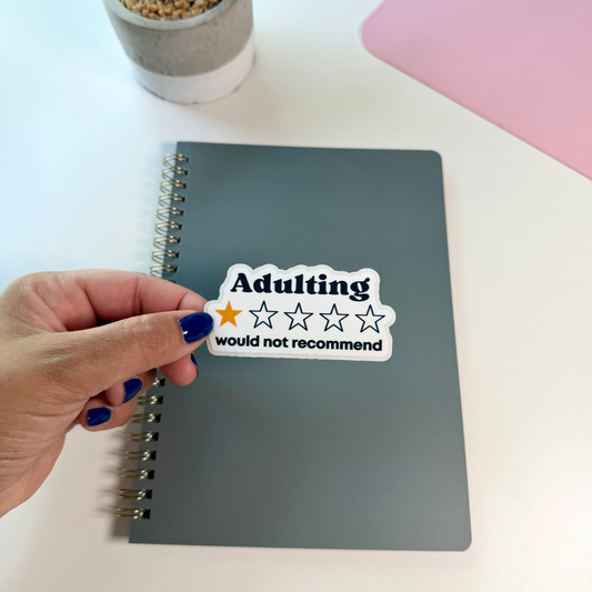 A hand with blue nail polish holds a white sticker that reads 'Adulting would not recommend' with a one-star rating, placed over a gray spiral notebook on a white desk with a small potted plant and a pink laptop edge in the background.