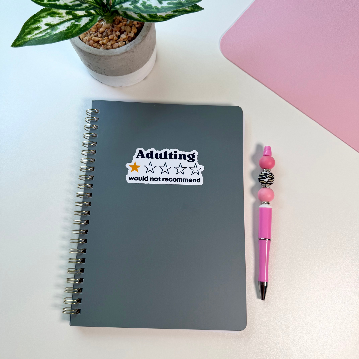 A dark gray spiral notebook with a sticker that reads 'Adulting would not recommend' is shown on a white desk next to a pink pen and a potted plant.