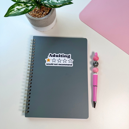 A dark gray spiral notebook with a sticker that reads 'Adulting would not recommend' is shown on a white desk next to a pink pen and a potted plant.