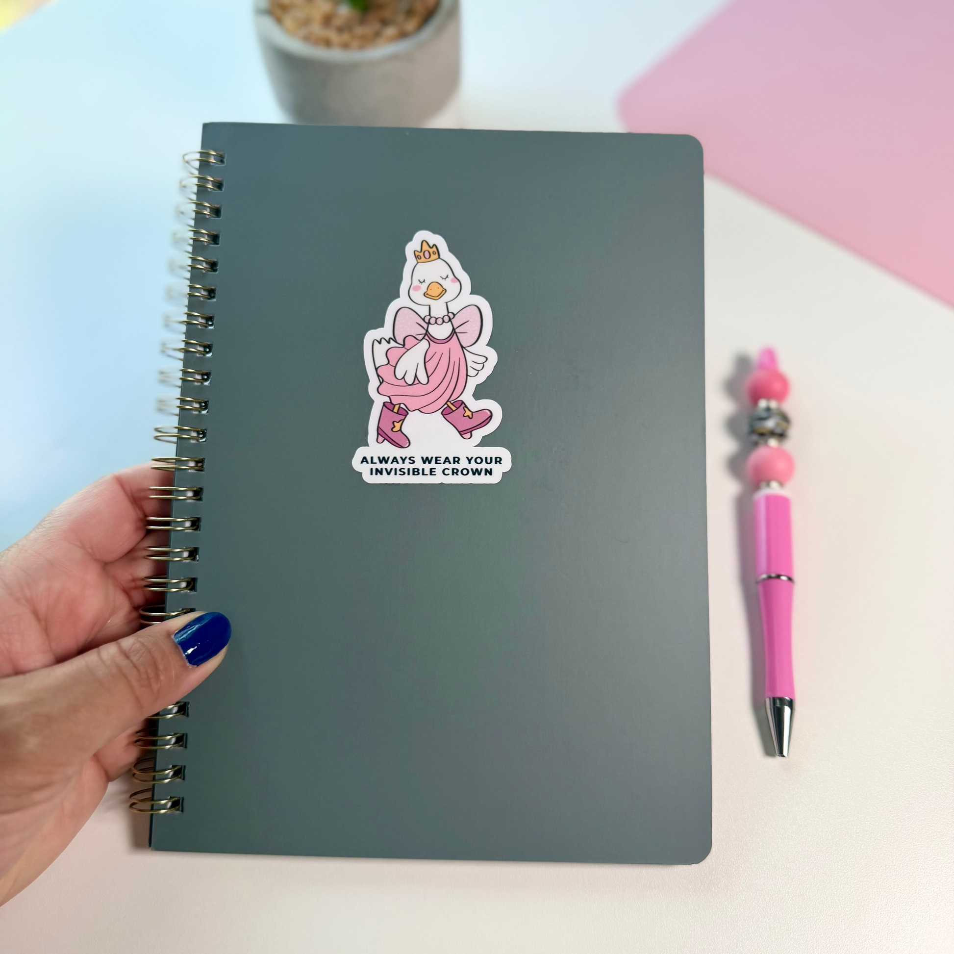 A dark gray spiral notebook with a pink cartoon duck sticker on the cover, held by a hand with blue nail polish next to a pink pen on a light desk with a potted plant in the background.