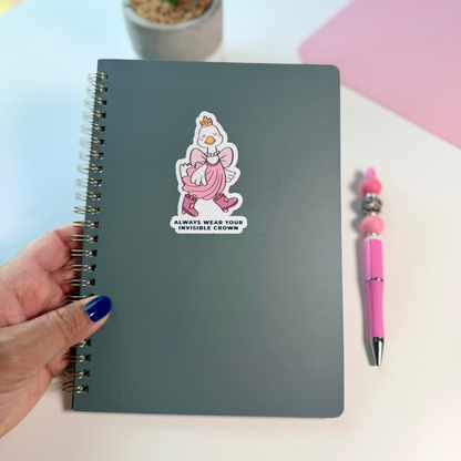 A dark gray spiral notebook with a pink cartoon duck sticker on the cover, held by a hand with blue nail polish next to a pink pen on a light desk with a potted plant in the background.