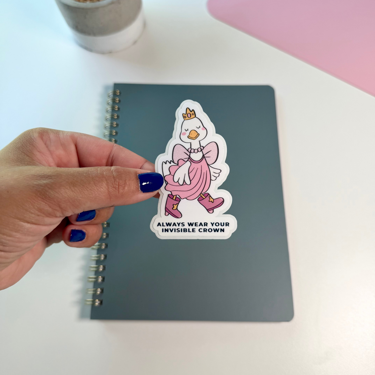 A hand with dark blue nail polish holds a die-cut sticker featuring a cartoon duck in a pink dress and crown above the text 'ALWAYS WEAR YOUR INVISIBLE CROWN'. The sticker is placed on a dark gray spiral notebook on a white desk, with a small potted plant and a pink laptop visible in the background.