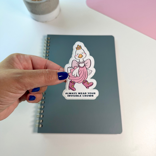 A hand with dark blue nail polish holds a die-cut sticker featuring a cartoon duck in a pink dress and crown above the text 'ALWAYS WEAR YOUR INVISIBLE CROWN'. The sticker is placed on a dark gray spiral notebook on a white desk, with a small potted plant and a pink laptop visible in the background.
