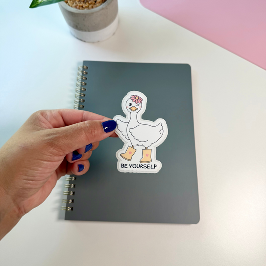 A hand with blue nail polish peels a die-cut sticker from a gray spiral notebook. The sticker features a cartoon white duck with Beige boots and a pink flower crown above the text 'BE YOURSELF'.