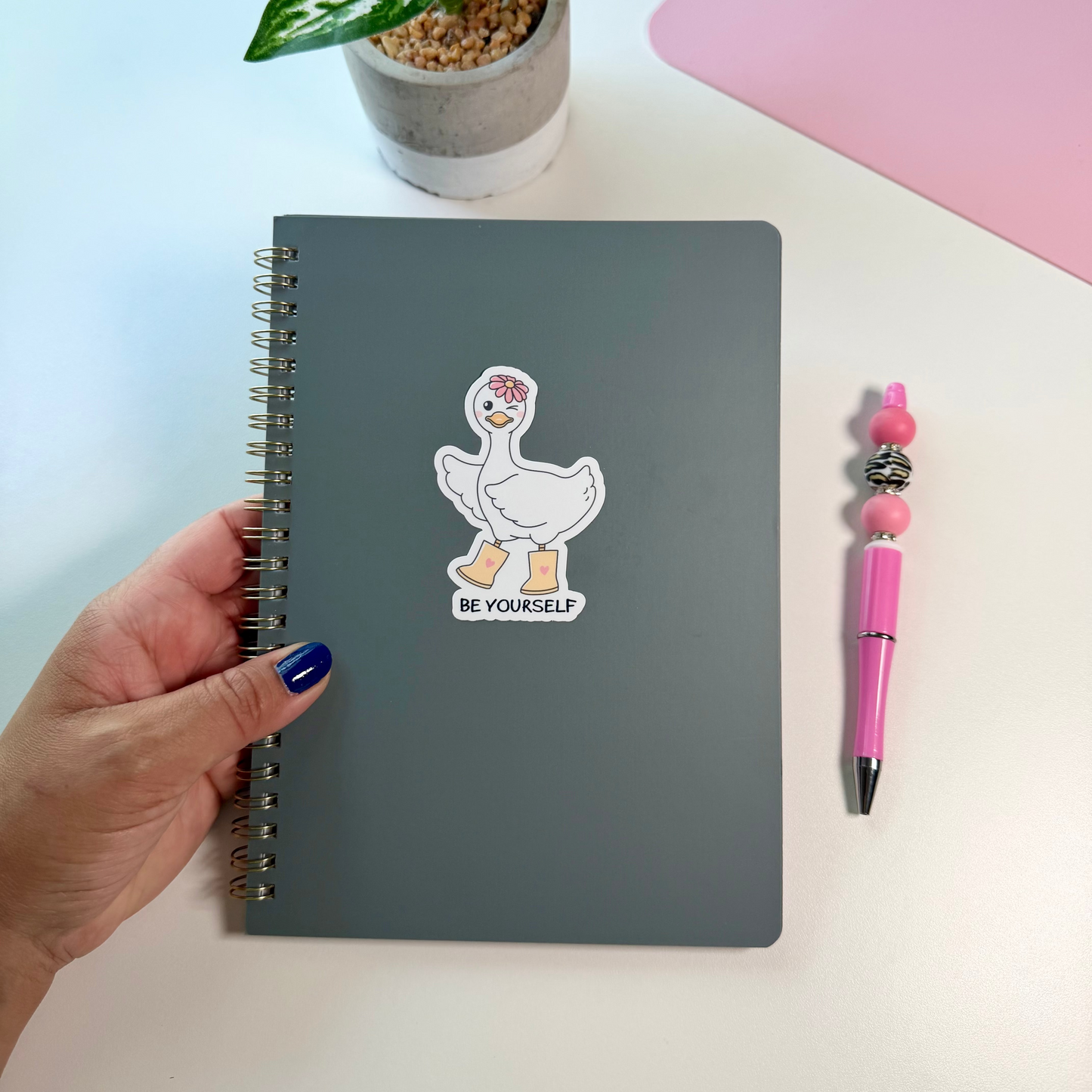 A hand with blue nail polish holds a dark gray spiral notebook featuring a white duck sticker with the text 'BE YOURSELF'. The notebook is displayed on a white desk alongside a pink pen and a small potted plant.