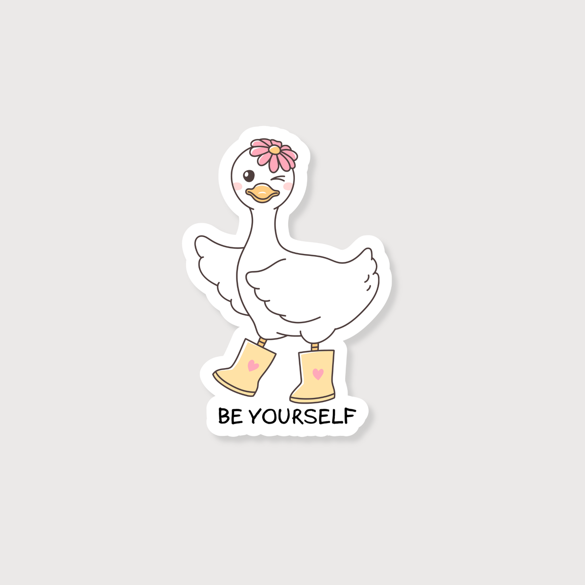 A die-cut sticker featuring a cute white cartoon goose wearing yellow rain boots with pink hearts, a pink flower on its head, and the text 'BE YOURSELF' below, shown on a light gray background.
