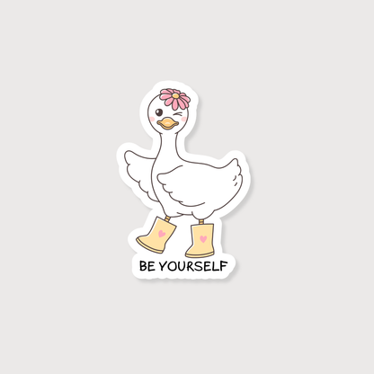 A die-cut sticker featuring a cute white cartoon goose wearing yellow rain boots with pink hearts, a pink flower on its head, and the text 'BE YOURSELF' below, shown on a light gray background.