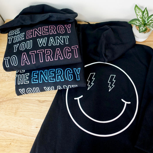 A stack of black hoodies laid out on a light wood surface against a white wall. The top hoodie has colorful text reading 'BE THE ENERGY YOU WANT TO ATTRACT,' the middle hoodie has blue text, and the front hoodie features a large white smiley face with lightning eyes.