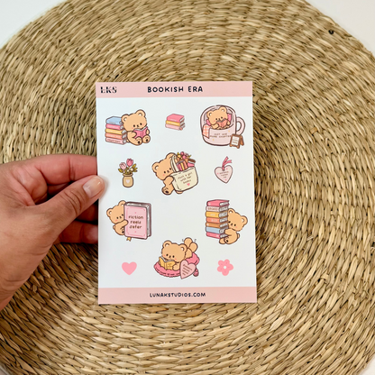 A hand holds a sheet of cute bookish-themed stickers against a white background with a round woven placemat underneath.