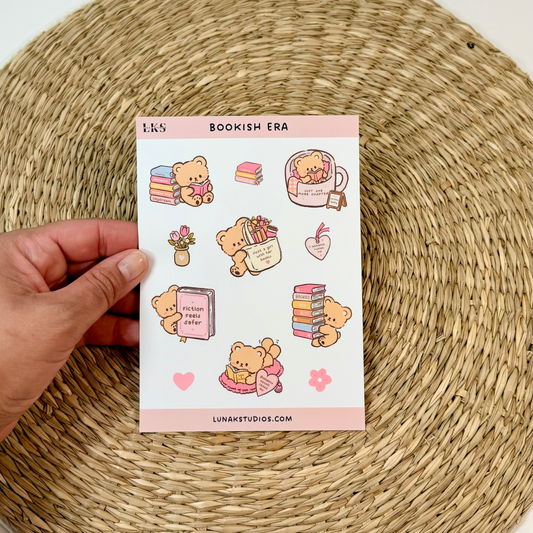 A hand holds a sheet of cute bookish-themed stickers against a white background with a round woven placemat underneath.