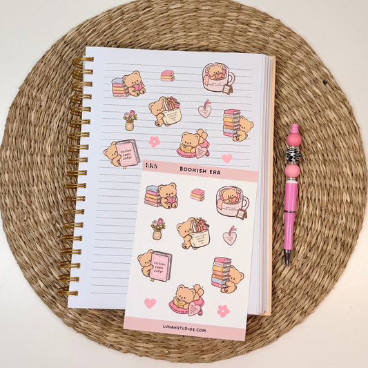 A spiral-bound planner sticker sheet and a matching pink pen arranged on a round woven placemat over a white tabletop. The planner stickers feature cute teddy bear illustrations with books, hearts, and small decorative motifs.