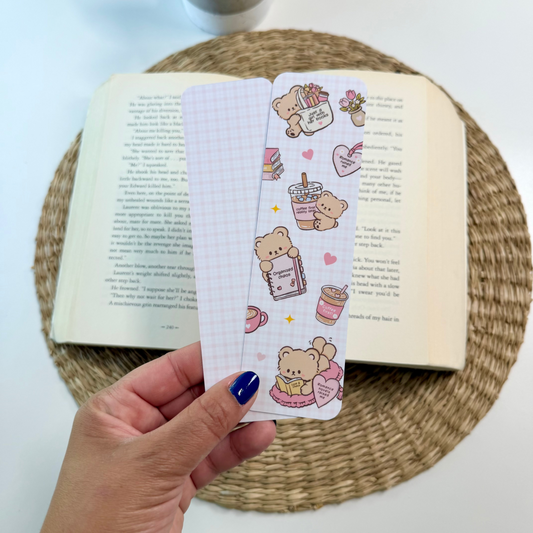 A hand with blue nail polish holds two cute illustrated bookmarks over an open book on a woven placemat. The bookmarks feature pastel pink and white designs with cartoon bears, coffee cups, hearts, and small food illustrations.