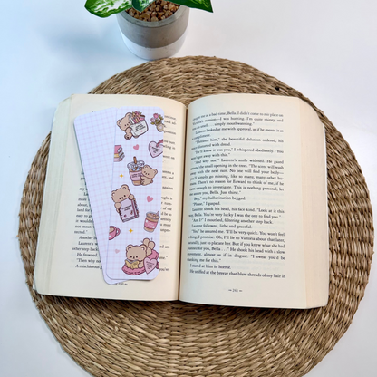 An open book resting on a round woven placemat, with a decorative pastel planner page featuring cute teddy bear illustrations placed on the left page. A small potted plant is visible in the background on a white surface.