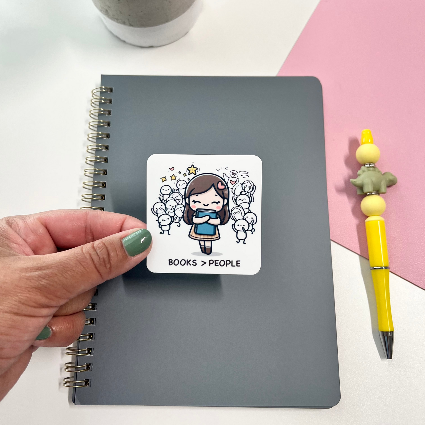 Person holding a sticker with a character design on a notebook with a plant and pen in the background.
