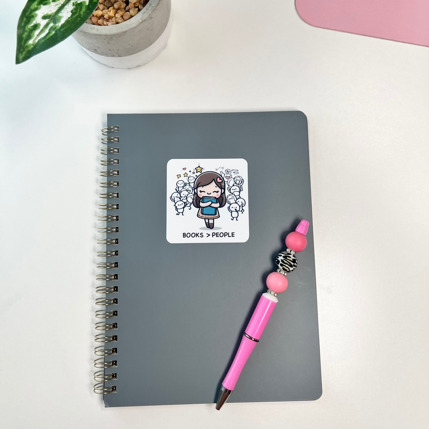 Gray notebook with a sticker, pink pen, and plant on a white surface