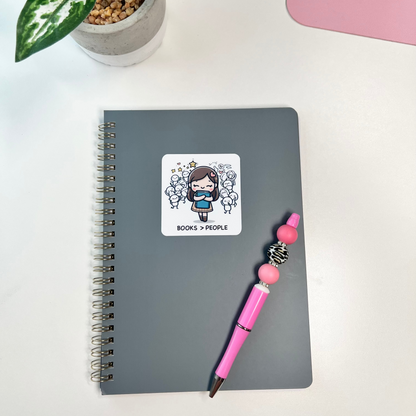 Gray notebook with a sticker, pink pen, and plant on a white surface