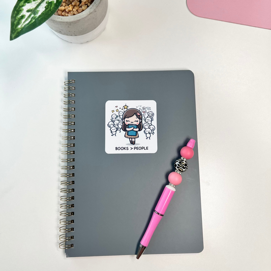 Gray notebook with a sticker, pink pen, and plant on a white surface