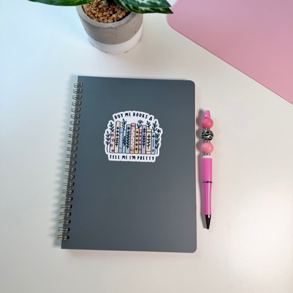 A dark gray spiral-bound notebook with a sticker on the cover, styled on a white desk alongside a pink pen and a potted plant.