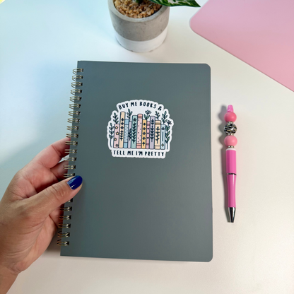 A hand with blue nail polish holds a dark gray spiral notebook with a colorful sticker that reads 'BUY ME BOOKS &  TELL ME I'M PRETTY'. The notebook sits on a white desk next to a pink pen and a small potted plant.