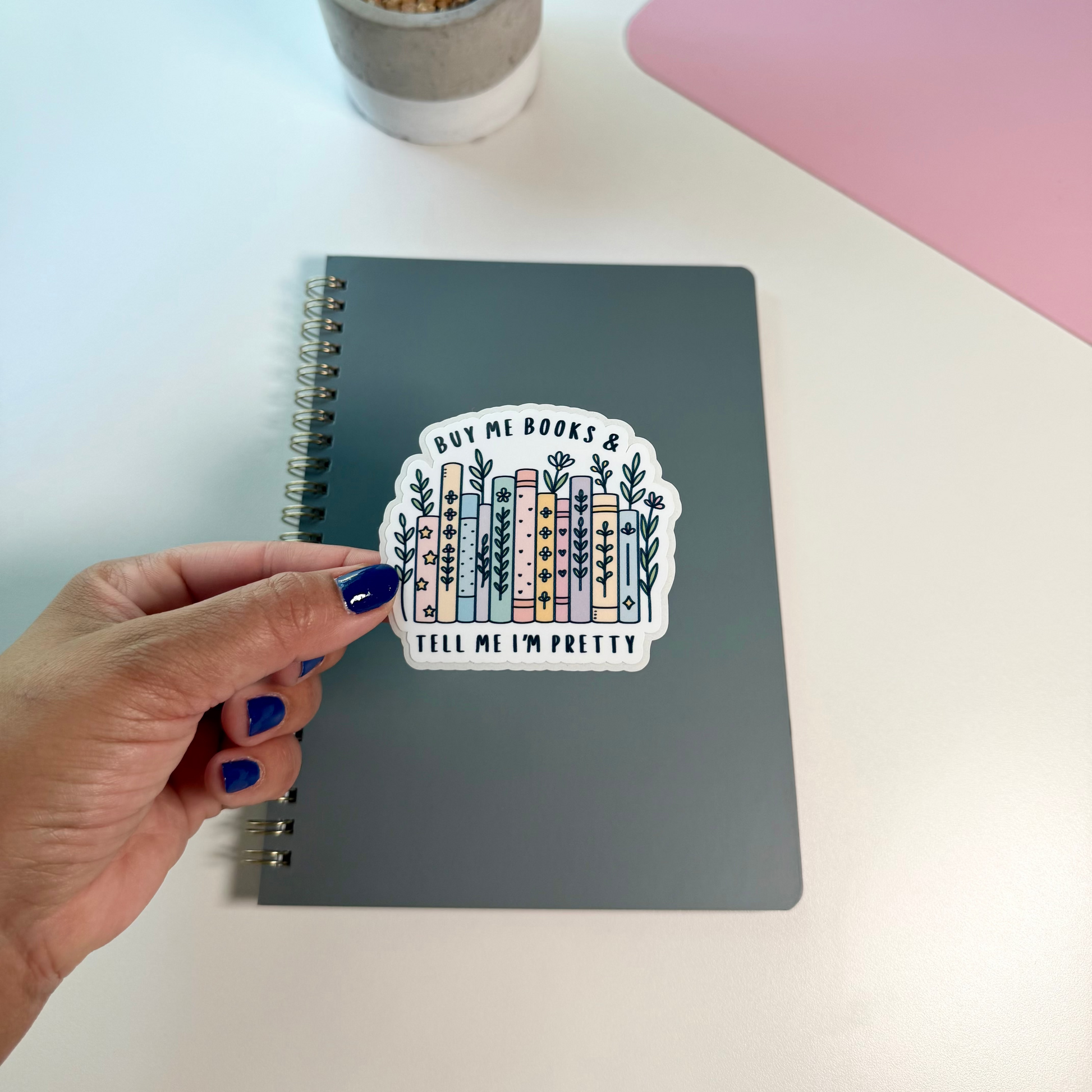 A hand with blue nail polish holds a die-cut sticker in front of a gray spiral notebook. The sticker features an illustration of colorful books with the text 'BUY ME BOOKS & TELL ME I'M PRETTY'.