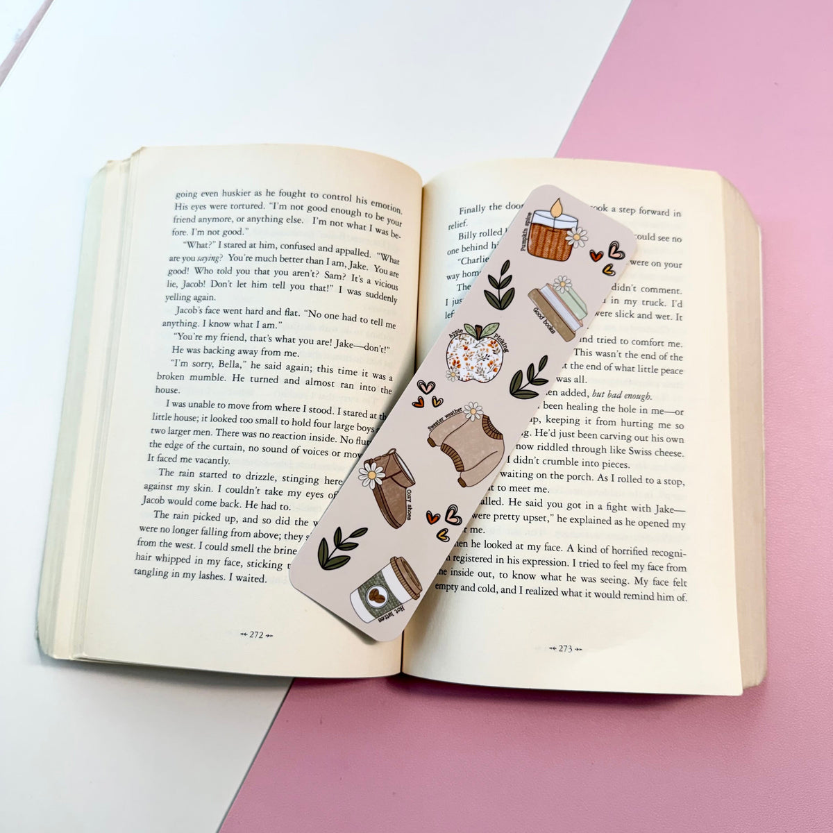A decorative bookmark with a cute illustration of a cozy kitchen scene, laid across an open book on a pink and white surface.