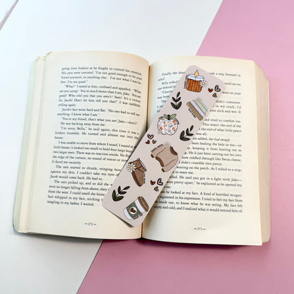 A decorative bookmark with a cute illustration of a cozy kitchen scene, laid across an open book on a pink and white surface.