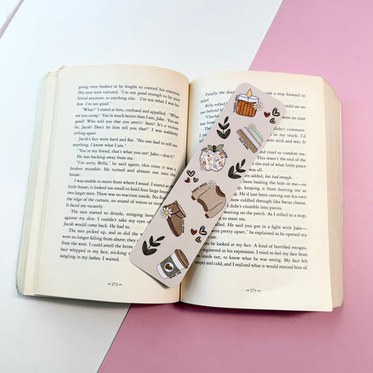 A decorative bookmark with a cute illustration of a cozy kitchen scene, laid across an open book on a pink and white surface.