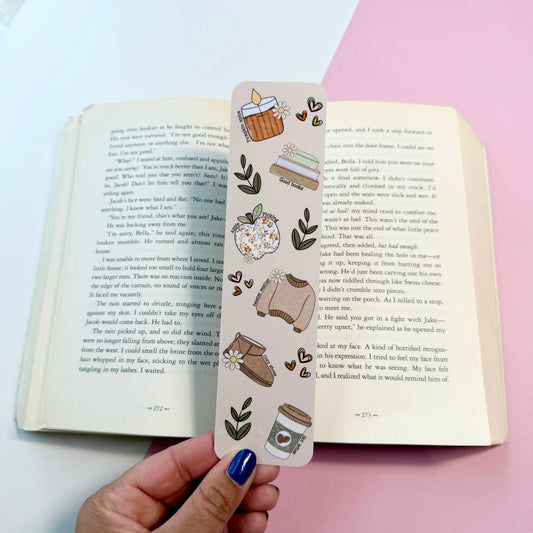 A hand with blue nail polish holds a decorative bookmark featuring cute illustrations of a coffee cup, books, leaves, and a small bag, positioned over an open book on a split white and pink background.