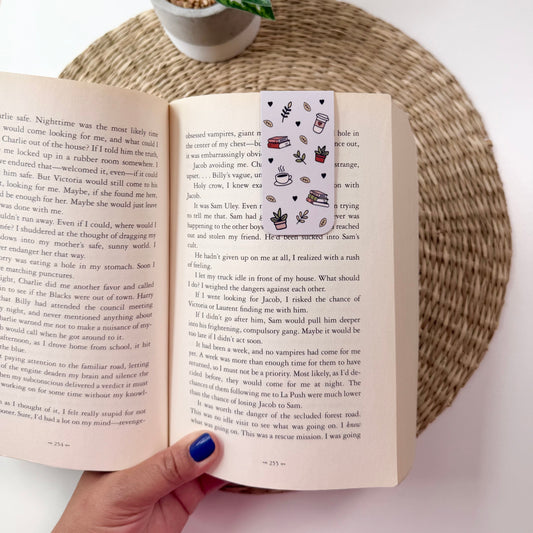 An open book with printed text and a small illustrated bookmark, held by a hand with blue nail polish. A woven placemat and a potted green plant are visible in the background.