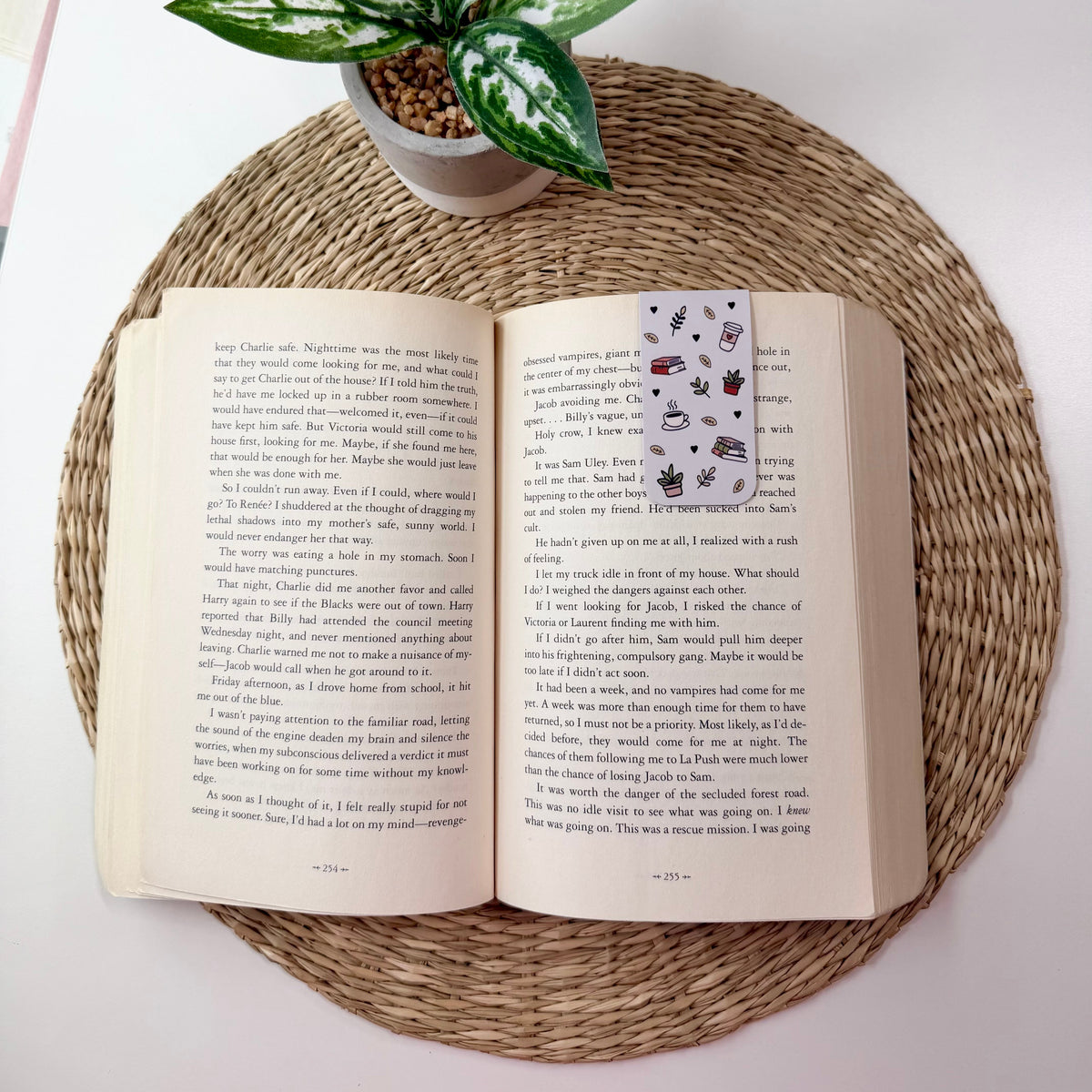 An open book spread on a round woven placemat, with a small potted green plant above it. The book pages are cream-colored with dense black text, and a small bookmark or card with a floral pattern is visible on the right page.