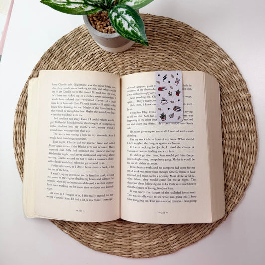 An open book spread on a round woven placemat, with a small potted green plant above it. The book pages are cream-colored with dense black text, and a small bookmark or card with a floral pattern is visible on the right page.