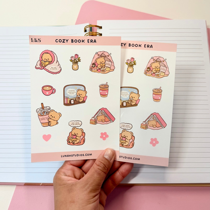 A hand holds two sticker sheets featuring cute cartoon illustrations themed around cozy books and drinks, set against a pink and white stationery background.