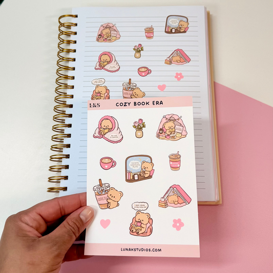 A hand holds a sheet of cute pink and beige sticker decals in front of an open spiral notebook on a white and pink surface.