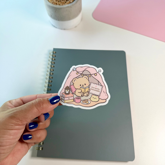 A hand with dark blue nail polish holds a die-cut sticker featuring a cute cartoon bear in a pink tent next to a gray spiral notebook on a white desk, with a potted plant and a pink desk mat visible in the background.