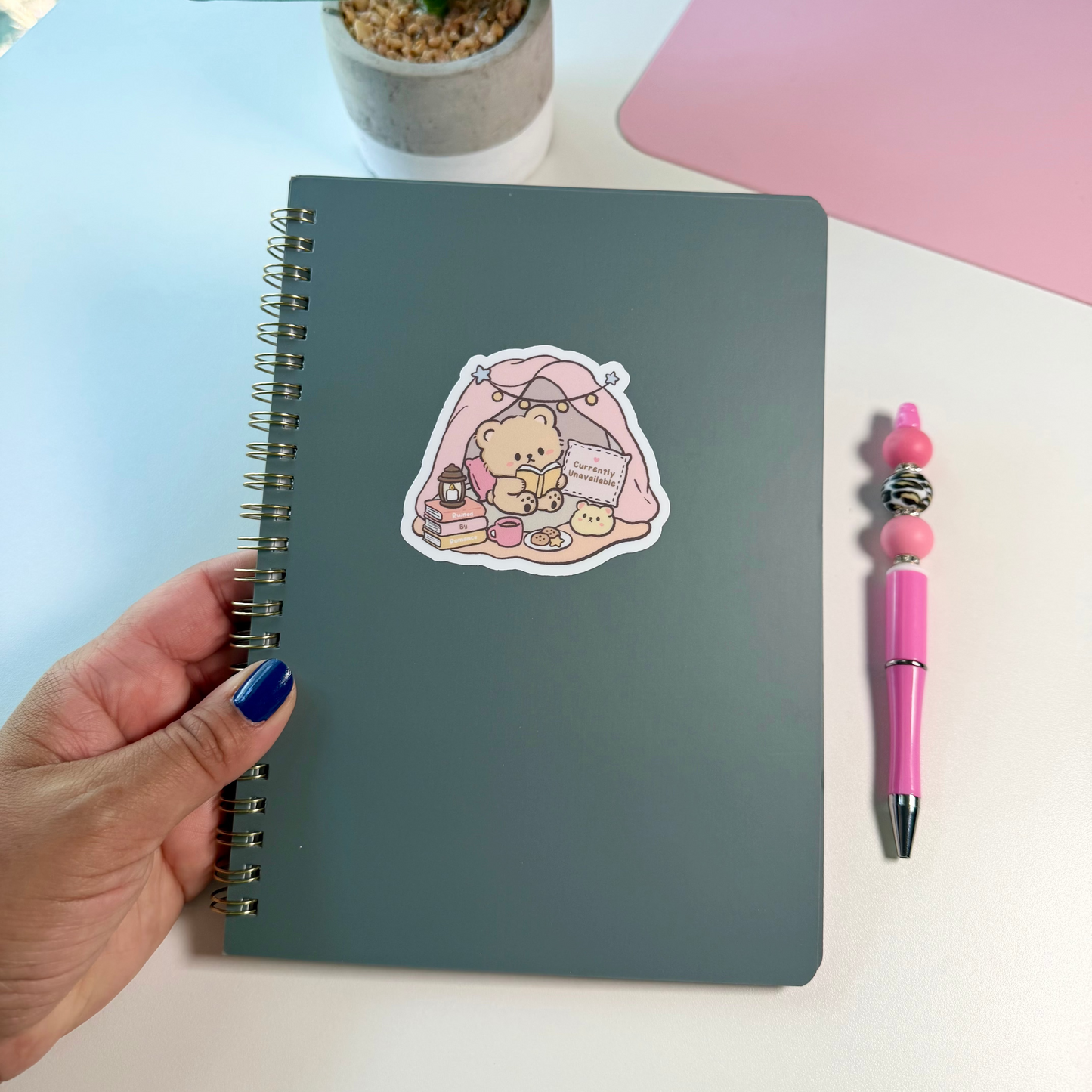 A dark gray spiral notebook with a cute sticker on the cover, held by a hand with blue nail polish. A pink pen and a small potted plant sit on a white desk next to a pink surface in the background.