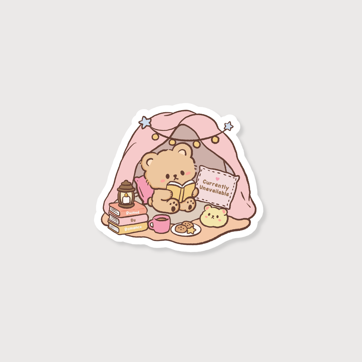 A die-cut sticker featuring a cute cartoon teddy bear sitting inside a pink blanket tent, reading a book with a mug, snacks, and a sign that says 'currently unavailable' on a light gray background.