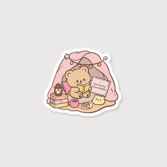 A die-cut sticker featuring a cute cartoon teddy bear sitting inside a pink blanket tent, reading a book with a mug, snacks, and a sign that says 'currently unavailable' on a light gray background.
