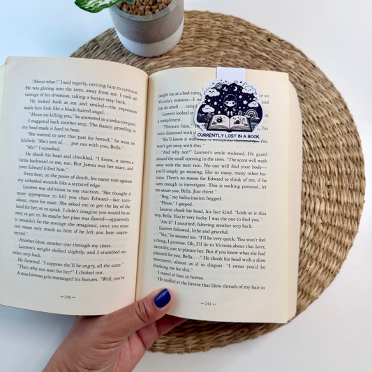 An open vintage book with printed text and a decorative crest on the right page, held by a hand with blue nail polish. The book is placed on a woven placemat next to a small potted plant against a white background.