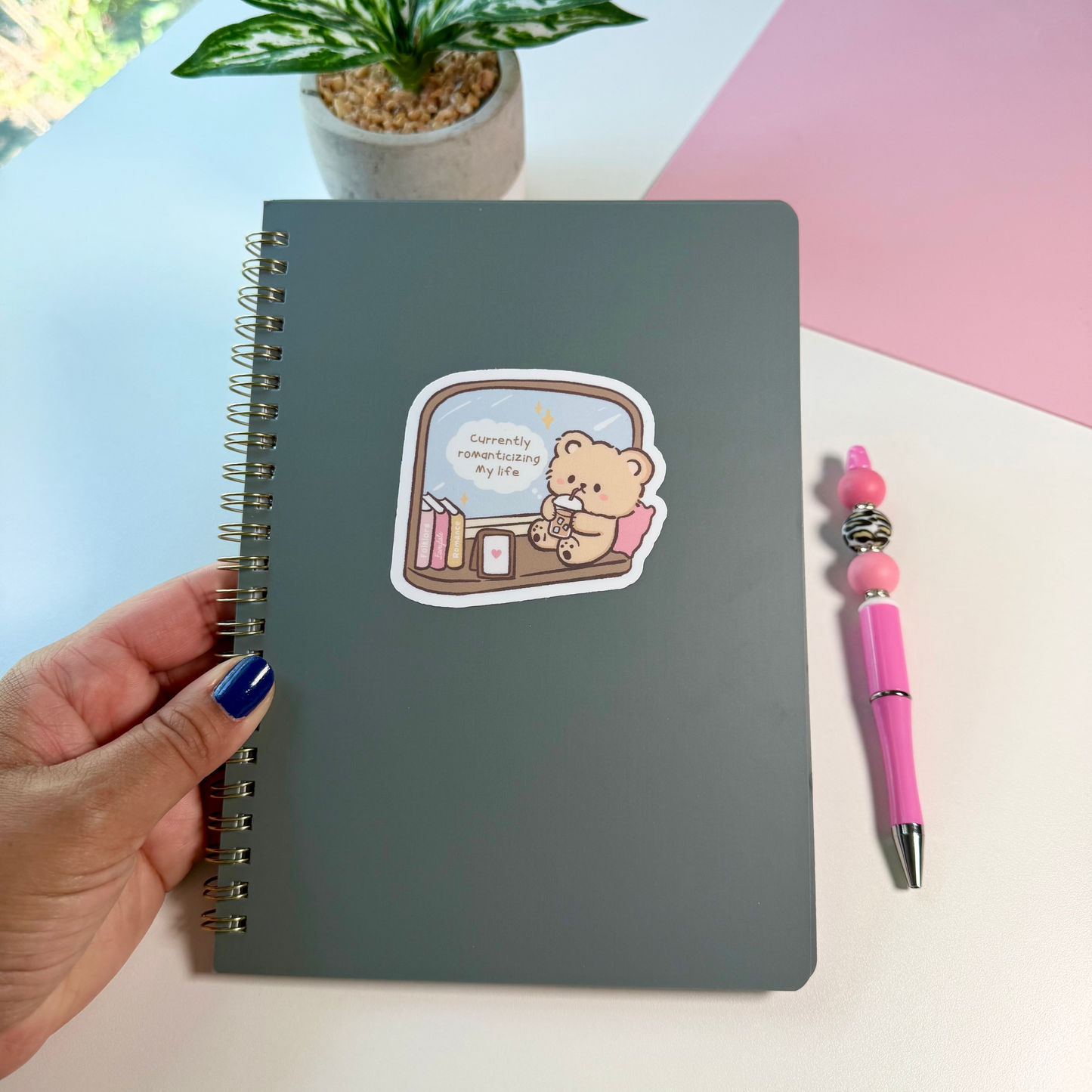 A dark gray spiral-bound notebook with a cute cartoon bear sticker on the cover, held by a hand with blue nail polish next to a pink pen on a white desk with a pink background.