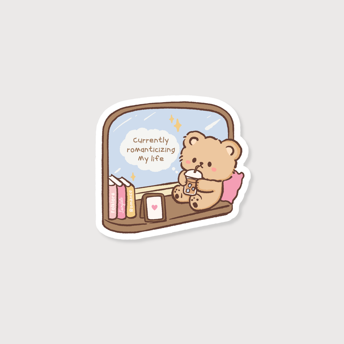 A cute cartoon bear sticker or decal showing a small tan bear sitting by a window, holding a phone, with books and a phone case on the windowsill and a speech bubble that reads 'Currently romanticizing my life'.